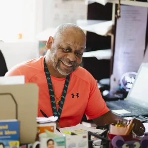Chester smiling in a bright orange shirt sitting at a busy desk. He is surrounded by office supplies, papers, and a laptop.