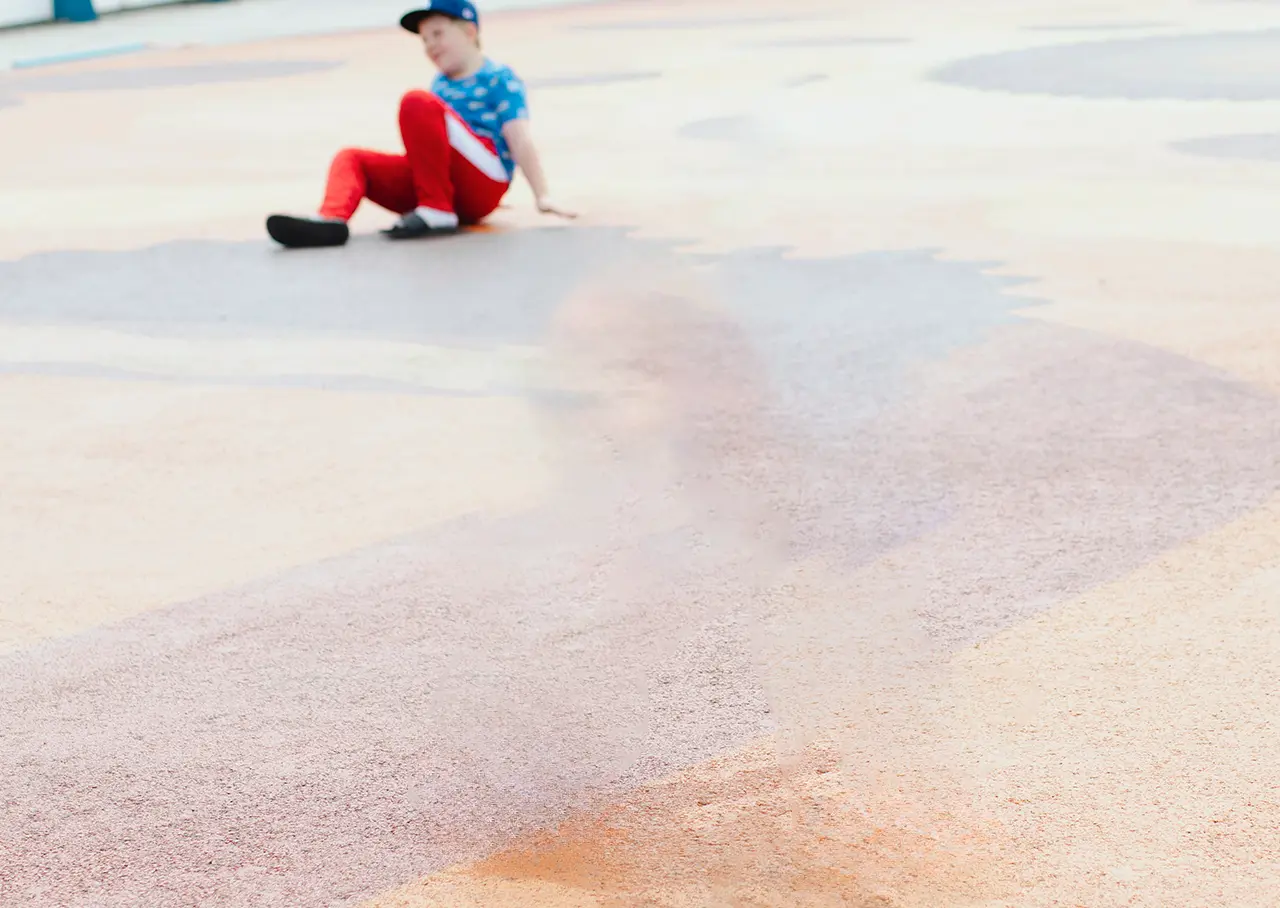 A young boy wearing a blue shirt, red pants, and a dark cap sits on the ground.