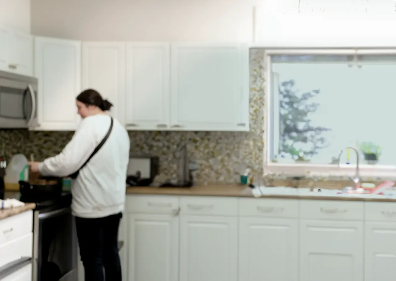 A person stands at a stove in a bright, modern kitchen with white cabinets and a mosaic tile backsplash.