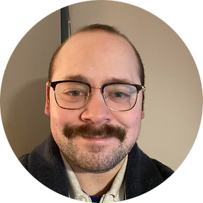A smiling man with glasses, a mustache, and short hair, wearing a dark jacket.
